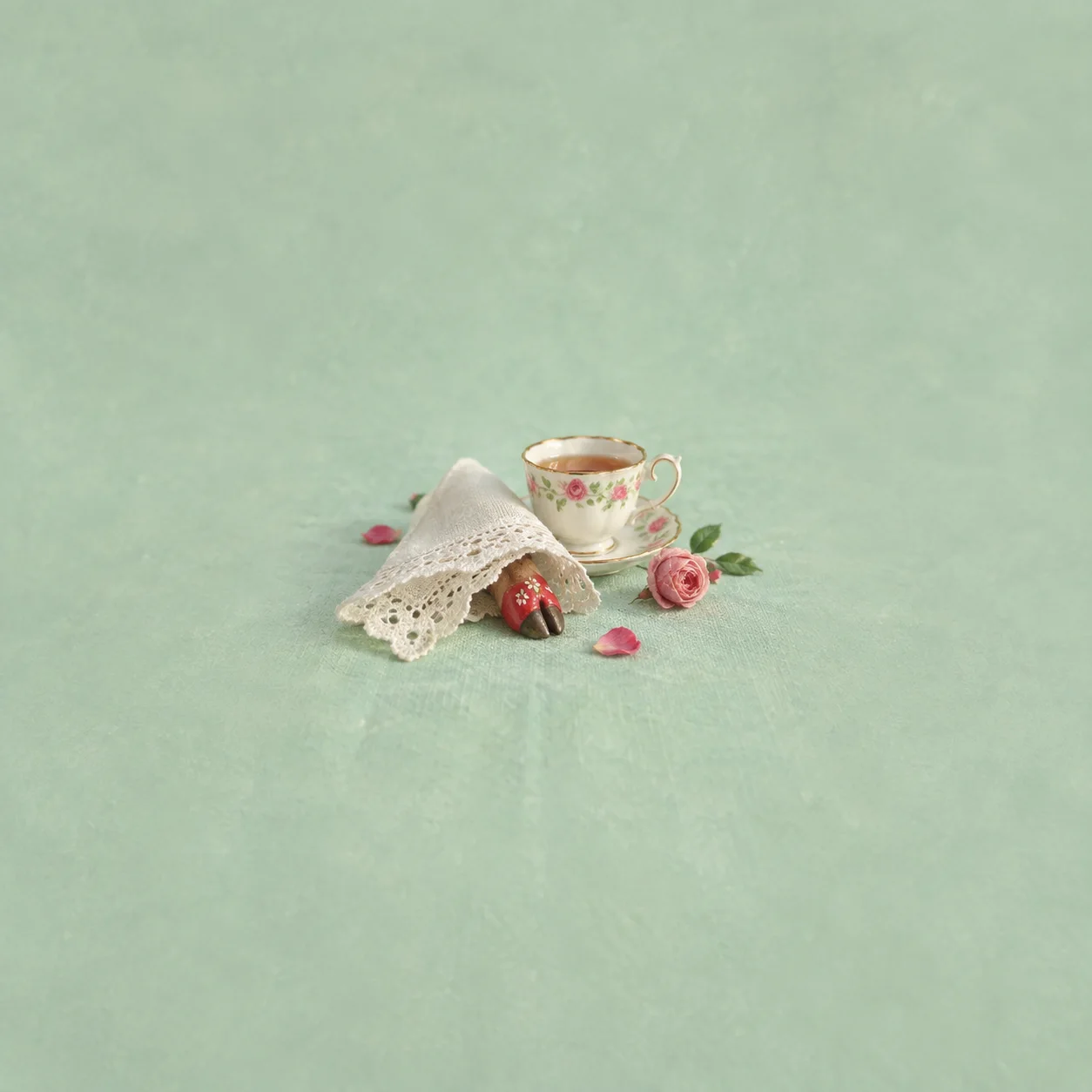 A floral teacup stands beside roses and a lace napkin with a small red cloven-hoof shape peeking out.